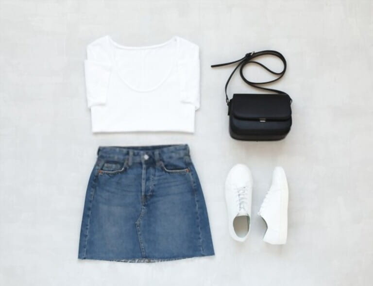 White t-shirt, blue denim mini skirt, small black crossbody bag, white sneakers on gray background. Aerial view of woman's casual dress. Lie flat, top view. White t-shirt, blue denim mini skirt, small black crossbody bag, white sneakers on gray background. Aerial view of woman's casual dress. Lie flat, top view.