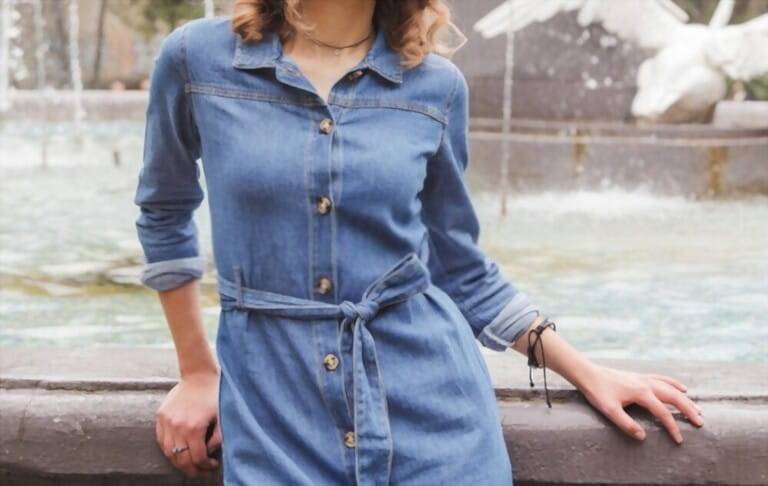 Slim woman in blue denim dress sitting on the fountain. Street look, summer outfit. Pretty female with curly hair posing in the city. 