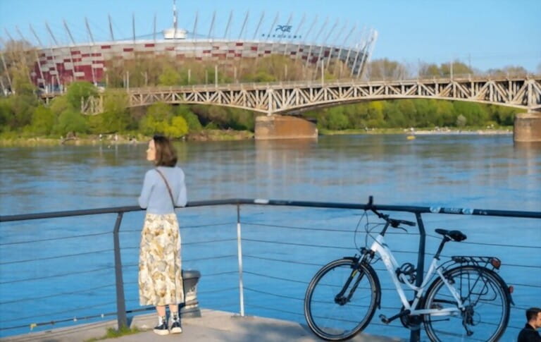 A girl in short jeans and a floral dress resting on a riverside square, next to a bicycle, a bridge, the National Stadium