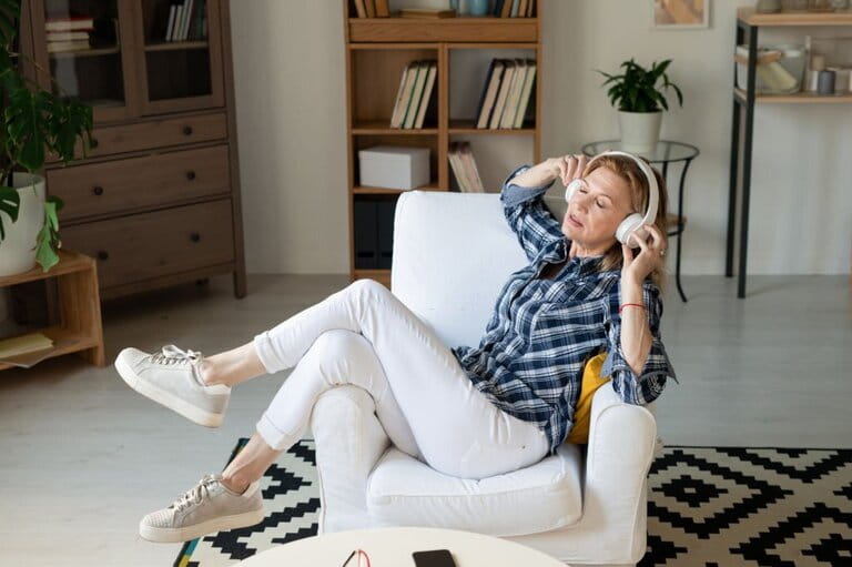 Comfortable mature woman in white jeans, gray sneakers and plaid shirt enjoying music sitting on sofa
