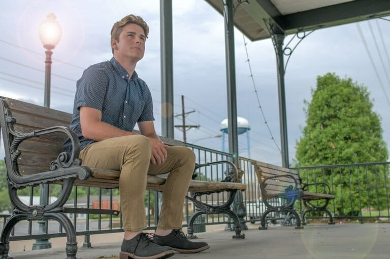 Cheerful young man sitting on a park bench in simple khaki pants, shirt and gray shoes Cheerful young man sitting on a park bench in simple khaki pants, shirt and gray shoes