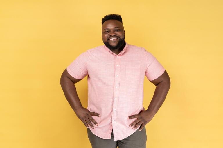 Portrait of happy satisfied man wearing pink shirt standing with hands on hips and looking at camera with toothy smile, expressing happiness. Indoor studio shot isolated on yellow background. Portrait of happy satisfied man wearing pink shirt standing with hands on hips and looking at camera with toothy smile, expressing happiness. Indoor studio shot isolated on yellow background.