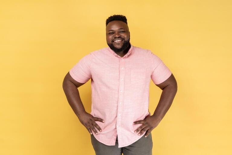 Portrait of happy satisfied man wearing pink shirt standing with hands on hips and looking at camera with toothy smile, expressing happiness. Indoor studio shot isolated on yellow background.
