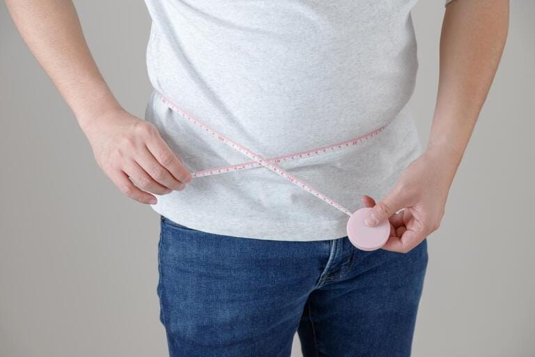 A man wearing a T-shirt and jeans is measuring his waist size, implying an image of health management and dieting.
