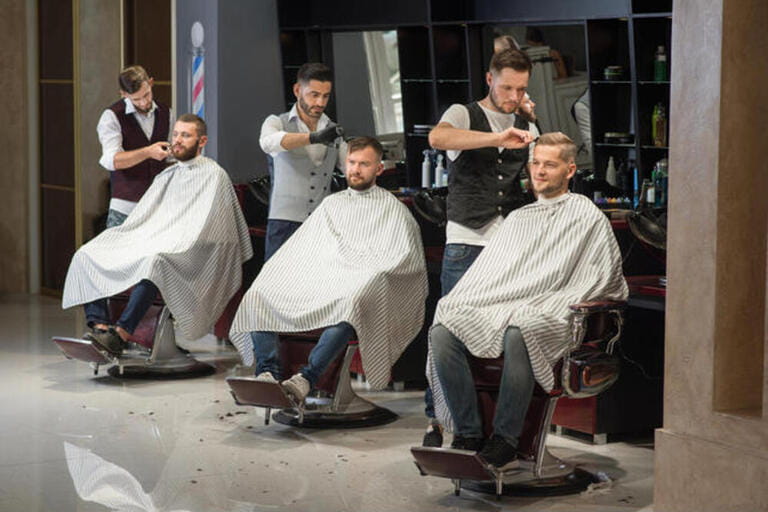 Process of styling and grooming men's haircuts in barbershop. Three professional and confident barbers standing and cutting hair of men. Male clients sitting in chairs and wearing haircut gowns Process of styling and grooming men's haircuts in barbershop. Three professional and confident barbers standing and cutting hair of men. Male clients sitting in chairs and wearing haircut gowns