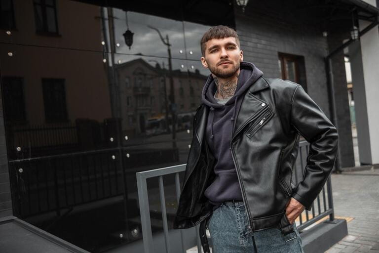 Cool stylish young hipster guy with hair, a beard, and a tattoo on his neck wearing fashion casual clothes with a leather rock jacket, hoodie, and jeans walks near a black modern building
