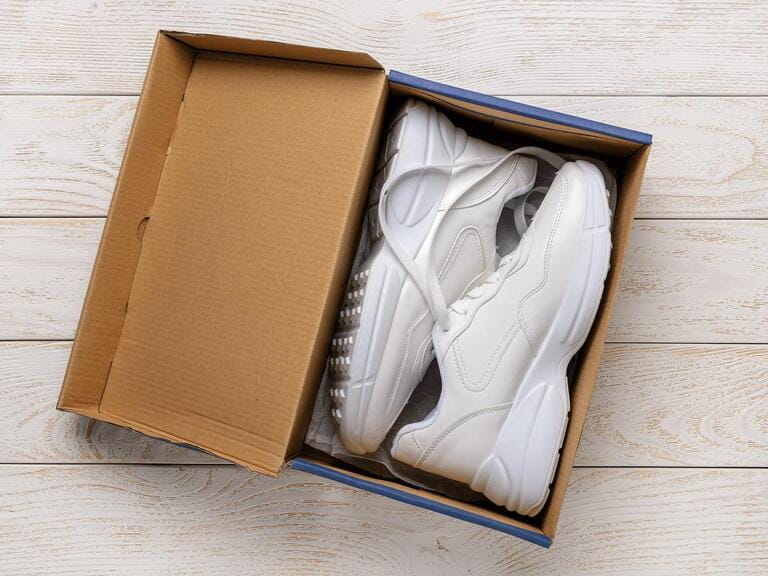 Pair of white chunky sole sneakers in a brown cardboard box on the white wood floor. Open box with new comfortable shoes for active lifestyle, fitness and sports. Top view.