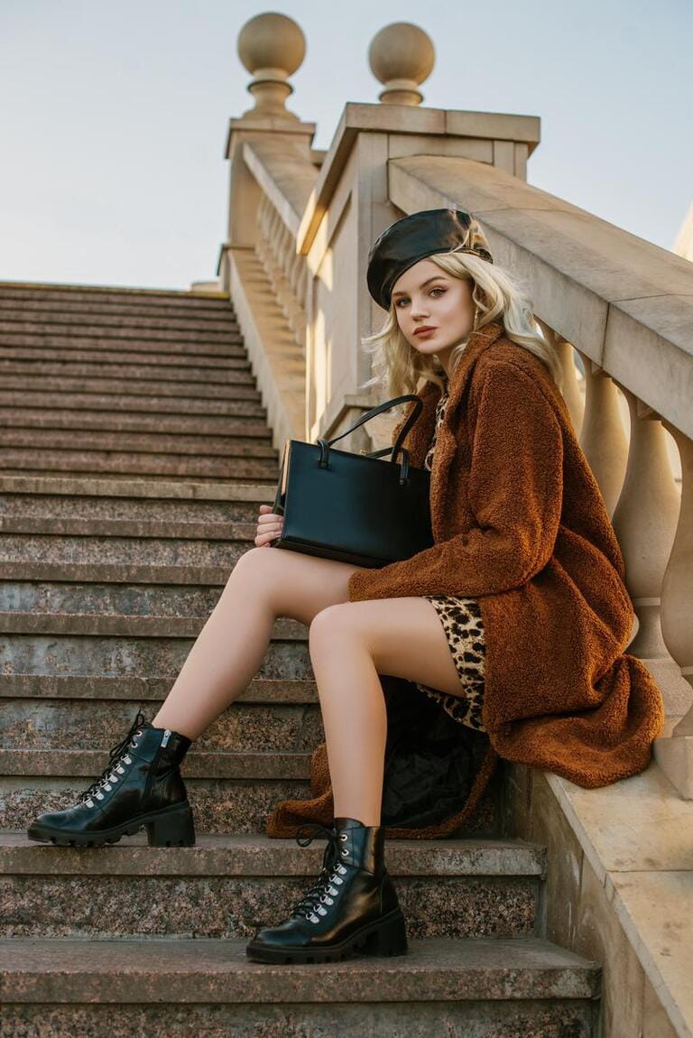 Outdoor full-length fashion portrait of young elegant woman wearing trendy brown faux fur coat, leather beret, lace up ankle boots, holding classic handbag, sitting, posing on stairs, in street Outdoor full-length fashion portrait of young elegant woman wearing trendy brown faux fur coat, leather beret, lace up ankle boots, holding classic handbag, sitting, posing on stairs, in street