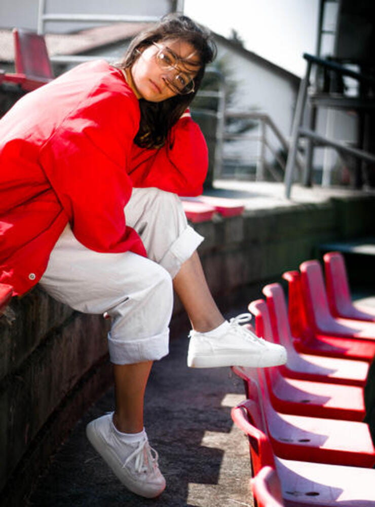 Young tennager girl model in the seat of a baseball stadium wearing red bomber jacket, hipster rounded glasses, white pants and sneakers