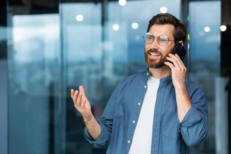 Successful smiling businessman in casual shirt talking on phone, male boss in glasses and beard near window inside modern office.
