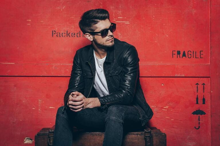 Gorgeous and stylish. Handsome young man in sunglasses holding hands tight and looking away while sitting on wooden chest against red background