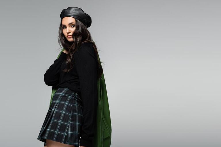 brunette woman in checkered skirt, black beret and green leather jacket posing isolated on grey brunette woman in checkered skirt, black beret and green leather jacket posing isolated on grey
