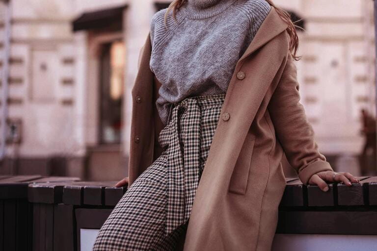 A woman in a fashionable outfit autumn brown coat, grey sweater and plaid pants on the city streets. Stylish female urban look. Film grain effect