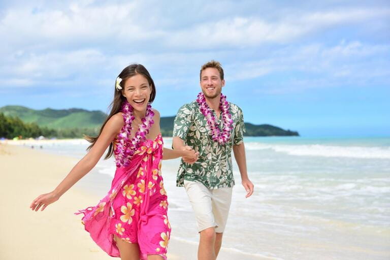 Happy couples have fun running on Hawaiian beach vacations in Hawaiian clothes wearing Aloha shirts and pink sarong sun dresses and floral leis for the traditional wedding or honeymoon concept.