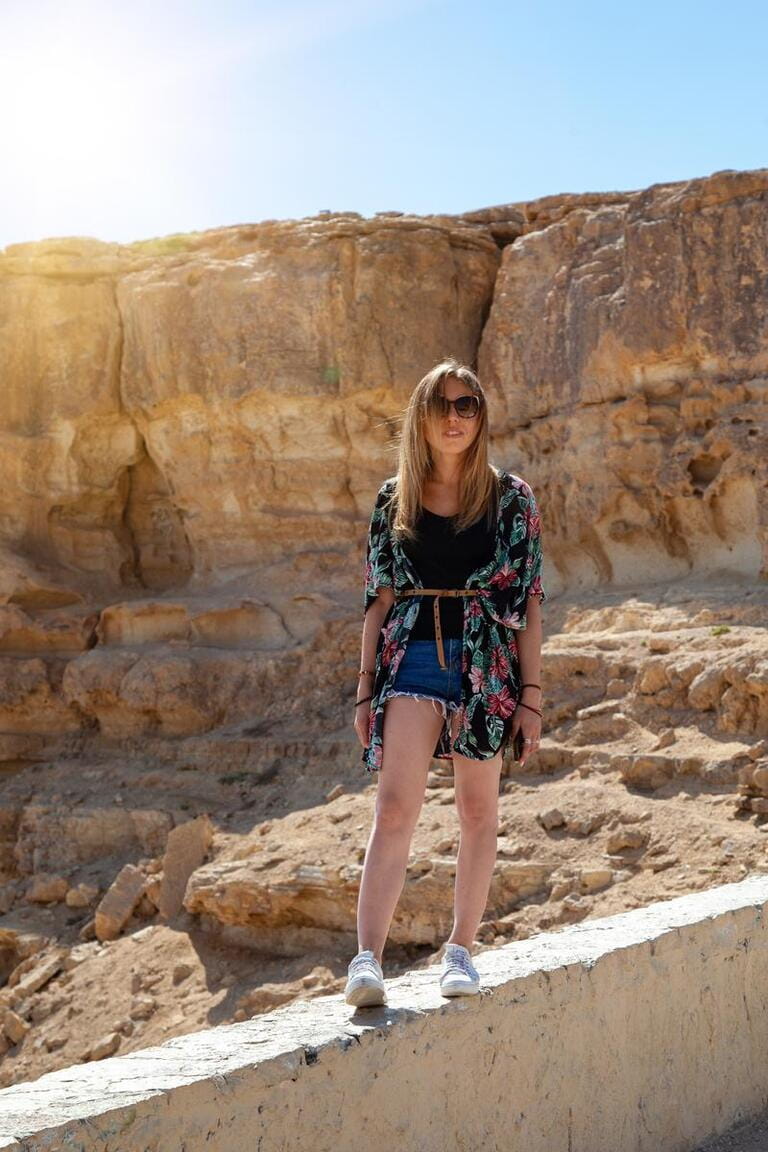 The model walks along the observation deck against the background of rocks in sunny weather. The girl is dressed like a tourist: sunglasses, blue shorts, a T-shirt and a Hawaiian shirt