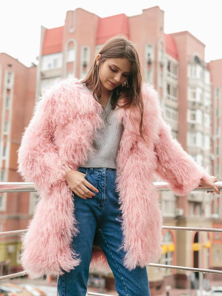 Young beautiful smiling woman with brunette wavy hair and makeup posing in pink lama fur coat at Milan street. City fashion. Trendy girl