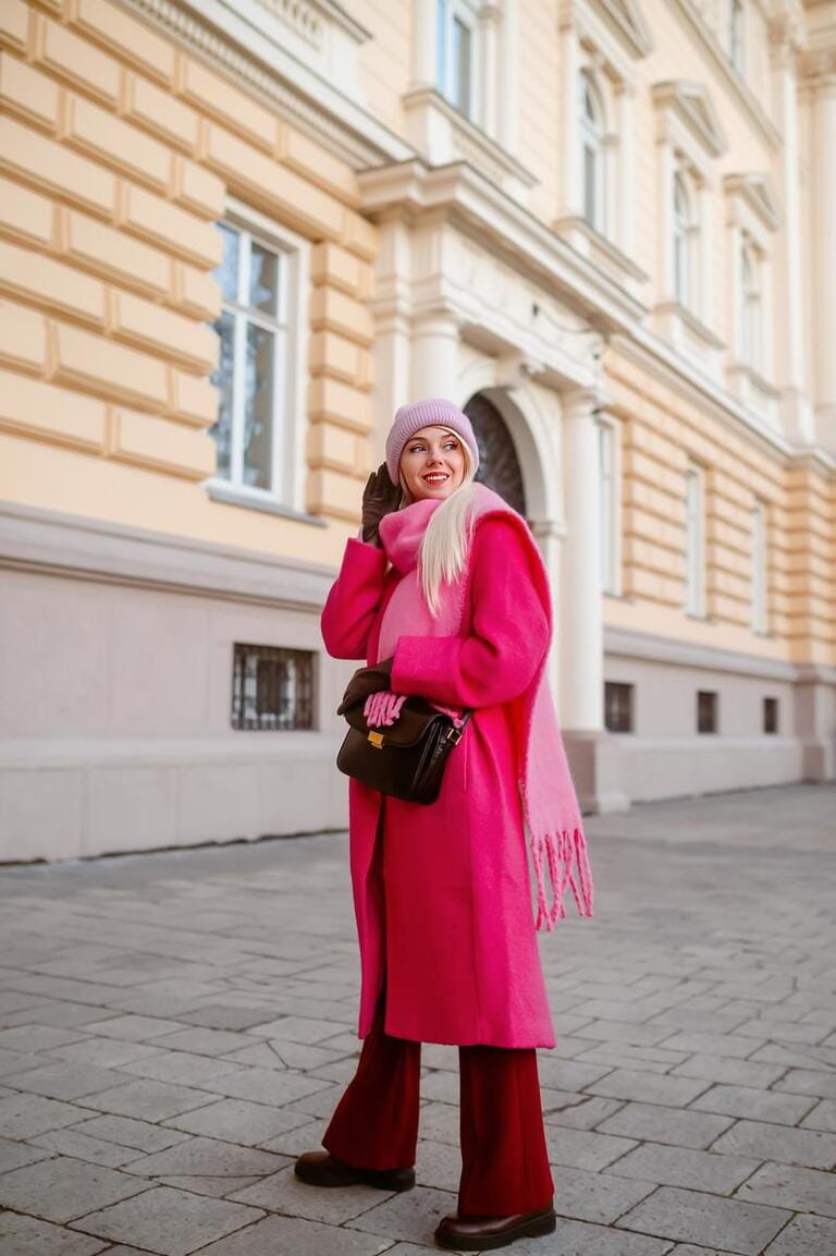 Happy smiling fashionable woman wearing trendy fuchsia color coat, pink beanie hat, fringed scarf, dark red flared trousers, leather boots, gloves. Street style conception.