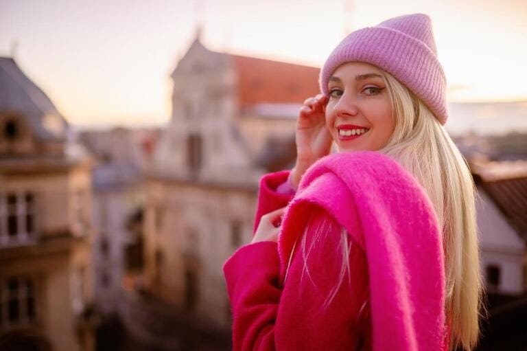 Happy smiling fashionable blonde woman wearing trendy knitted beanie hat, woolen scarf, coat, posing outdoor, with beautiful city view.