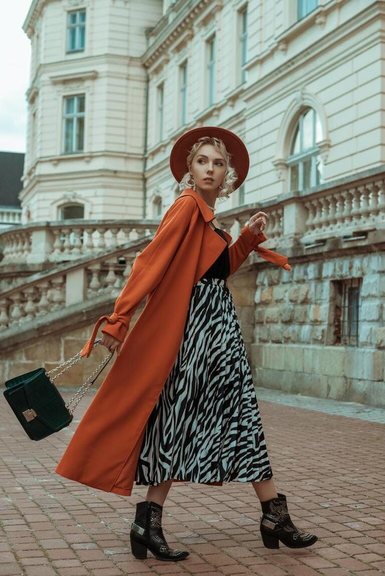Fashionable woman wearing long orange trench coat, hat, zebra print midi skirt, cowboy ankle boots, with green bag, walking in street of European city Fashionable woman wearing long orange trench coat, hat, zebra print midi skirt, cowboy ankle boots, with green bag, walking in street of European city