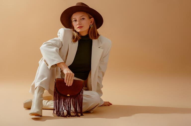 Fashionable confident woman wearing elegant white suit, hat, leather ankle boots, holding brown suede fringed bag, sitting, posing on beige background.