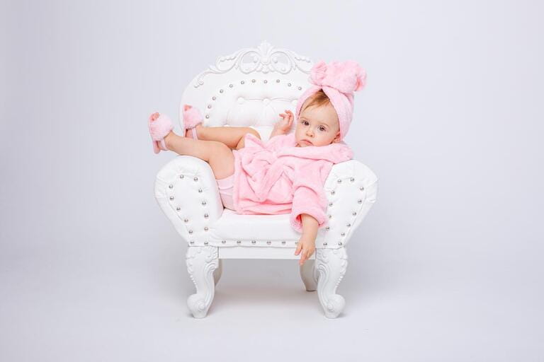 A cute little baby girl in a pink terry dressing gown with a chair and a mini dressing table with cosmetics is isolated on a white background, repeats the behavior of her mother A cute little baby girl in a pink terry dressing gown with a chair and a mini dressing table with cosmetics is isolated on a white background, repeats the behavior of her mother