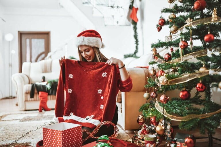 Cute woman receiving red reindeer sweater in Christmas gift box under the tree