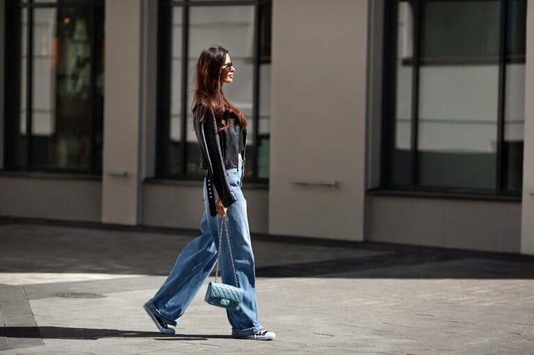 Beautiful young full-length brunette woman wearing black leather jacket and blue loose jeans, sneakers, sunglasses and small handbag with chain, walking street on sunny day Beautiful young full-length brunette woman wearing black leather jacket and blue loose jeans, sneakers, sunglasses and small handbag with chain, walking street on sunny day