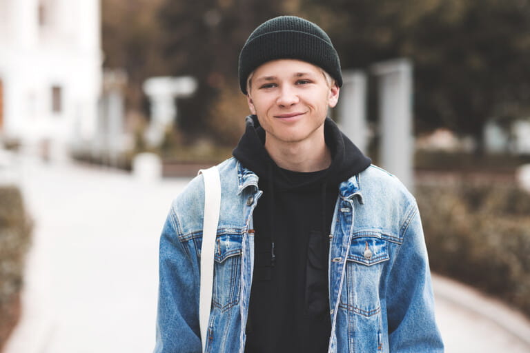 Smiling handsome teen boy 16-17 year old wearing denim jacket and knit hat over city background close up. Teenagerhood. Smiling handsome teen boy 16-17 year old wearing denim jacket and knit hat over city background close up. Teenagerhood.