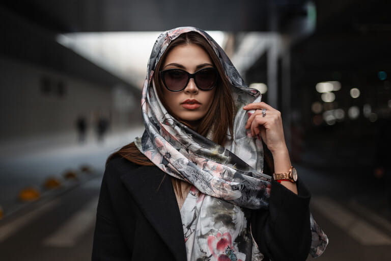 Elegant pretty young woman in fashionable clothes posing outdoors. Portrait beautiful girl in dark trendy sunglasses in a coat with a luxurious silk scarf on head in the city. Urban business lady.