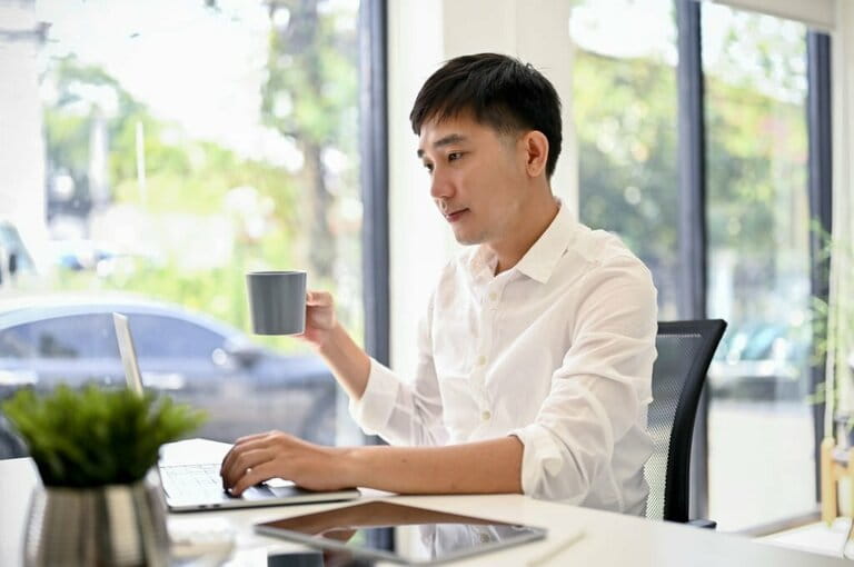 Handsome businessman using laptop and sipping coffee at desk in a modern office