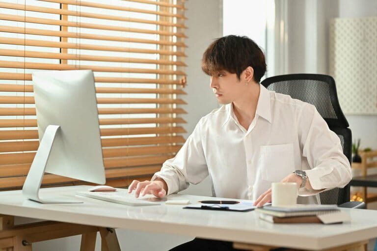 Concentrated young businessman wearing white shirt looking at computer screen, reviewing project in bright office interior. Concentrated young businessman wearing white shirt looking at computer screen, reviewing project in bright office interior.