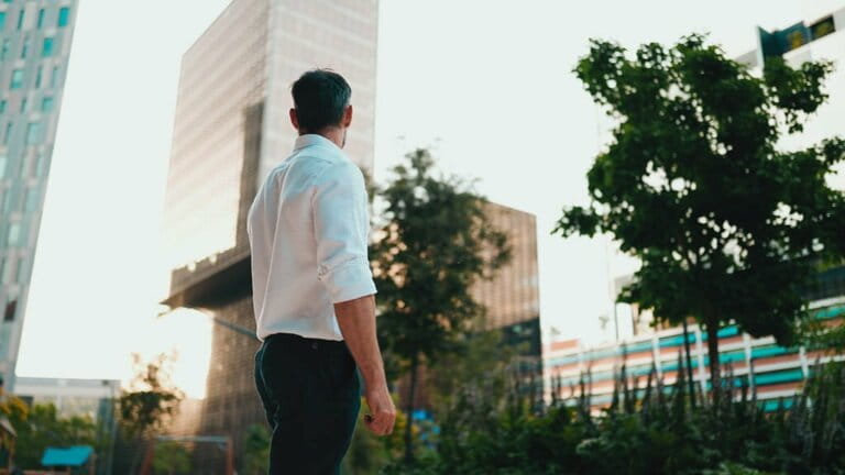 Mature businessman with neat beard wearing white shirt standing in financial district in city. Successful man looks around against the background of modern buildings