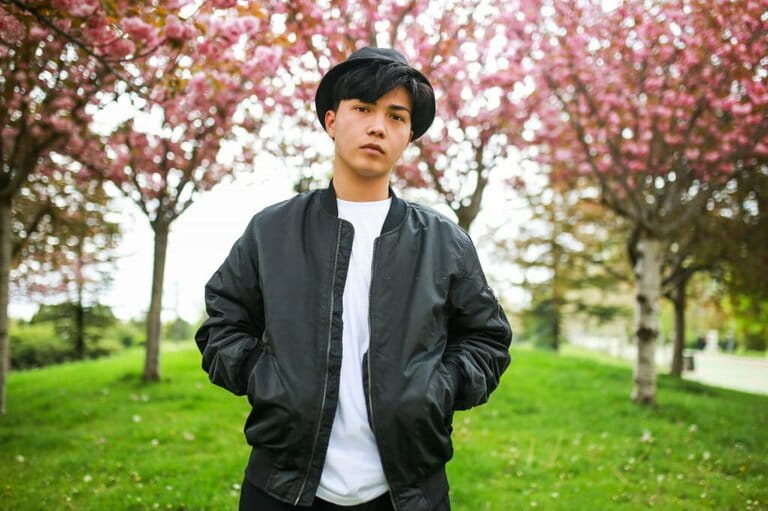 Portrait of young Asian man wearing bomber jacket and black hat standing in front of pink flowering tree in spring