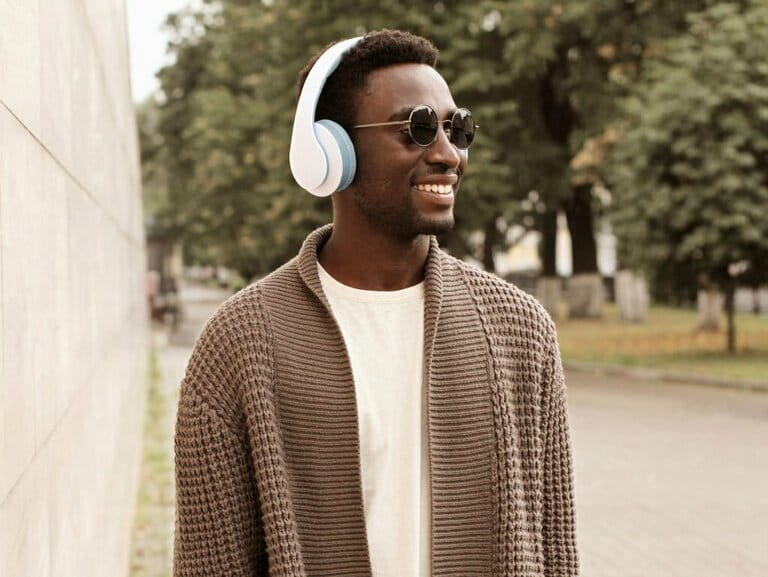 Portrait of young African man smiling happily in headphones listening to music wearing brown knitted cardigan in a city