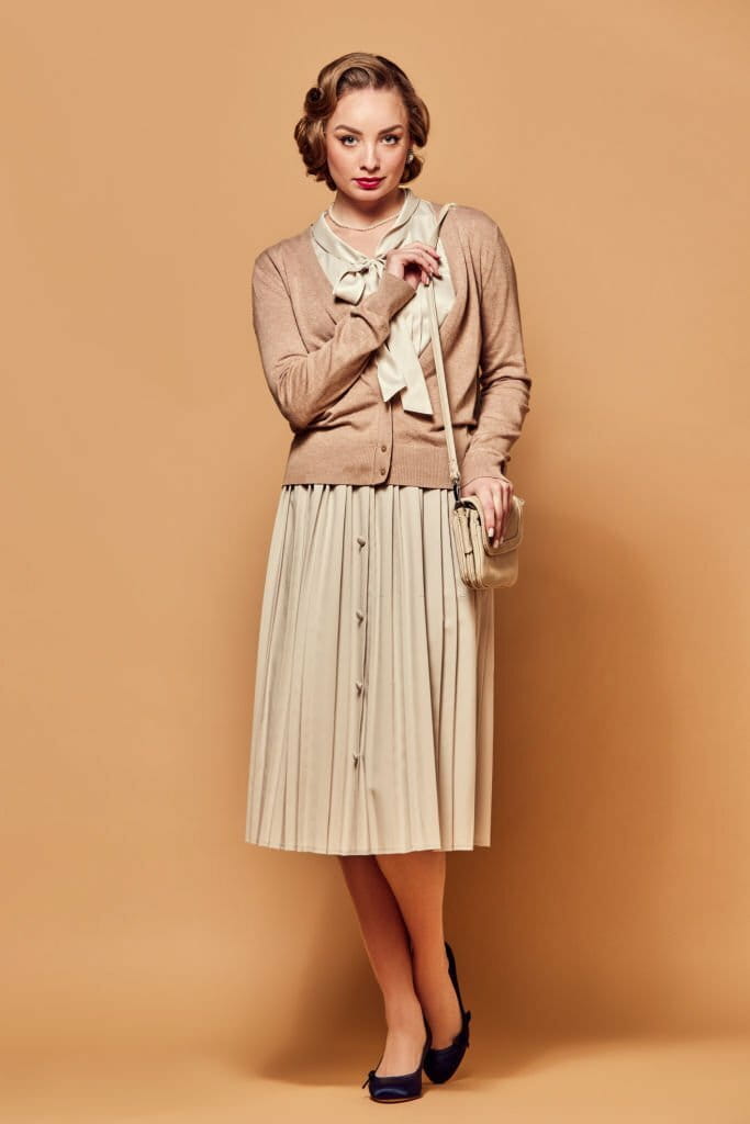 A beautiful woman wearing vintage clothes stands with a serious expression and looks at the camera against a beige background