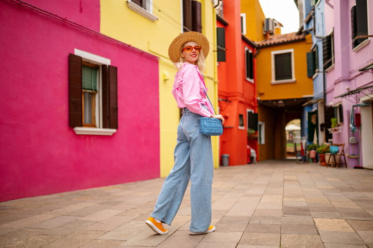 Happy smiling female traveler wearing stylish hat, glasses, pink shirt, wide leg trousers, walking, posing near colorful houses in street. Travel, tourism, vacation, fashion, lifestyle conception