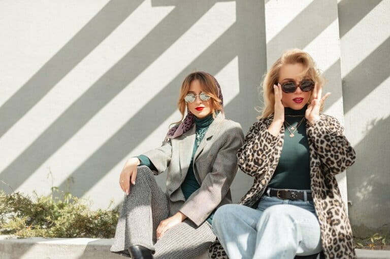 Two beautiful chic fashion girls in stylish clothes sitting on the street near a gray wall. Costumes in urban women's style Two beautiful chic fashion girls in stylish clothes sitting on the street near a gray wall. Costumes in urban women's style