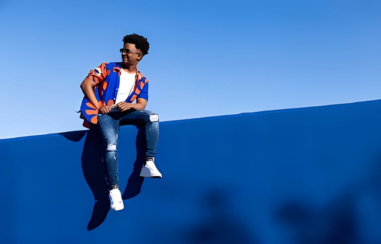 Full body of young african american male in trendy outfit and sneakers with eyeglasses sitting on blue concrete wall against blue sky in daylight Full body of young african american male in trendy outfit and sneakers with eyeglasses sitting on blue concrete wall against blue sky in daylight