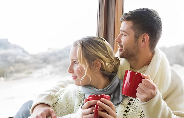 Young couple in winter clothing with coffee cups looking out through cabin window