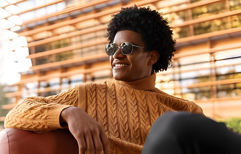 Trendy smiling african american male in knitted yellow sweater with legs crossed and hands together looking away on leather sofa in terrace near contemporary house Trendy smiling african american male in knitted yellow sweater with legs crossed and hands together looking away on leather sofa in terrace near contemporary house