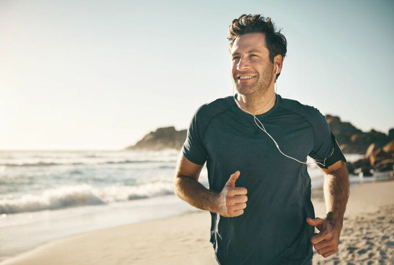 Active, fit and healthy man jogging on the beach while listening to music on headphones with beautiful outdoor clear sky and sea copy space