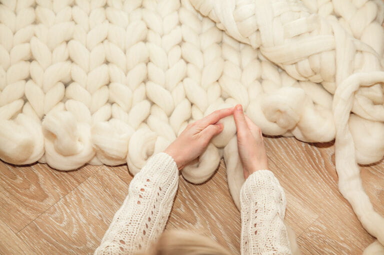 Woman's hands knitting a large merino wool plaid on wooden floor