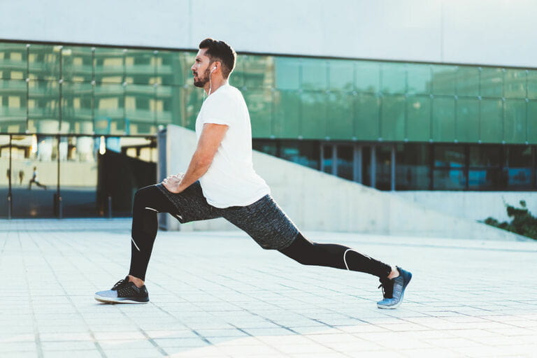 Flexible men in tracksuits warming up leg muscles during outdoor physical training, strong sports athletes listening to music in headphones during morning workouts Flexible men in tracksuits warming up leg muscles during outdoor physical training, strong sports athletes listening to music in headphones during morning workouts