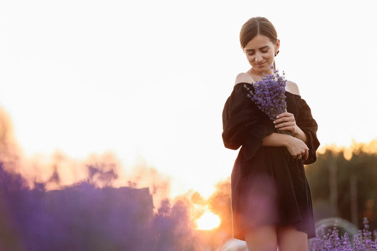 Romantic young woman with naturally beautiful healthy skin holding bouquet of flowers in lavender field. Beautiful girl wearing a black dress and bow in her hair.
