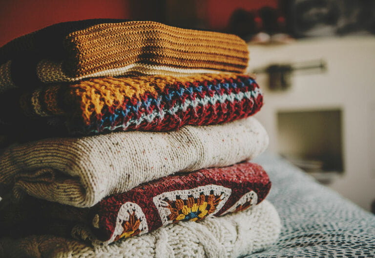 Cozy fall sweaters folded and stacked up on table by window at bedroom
