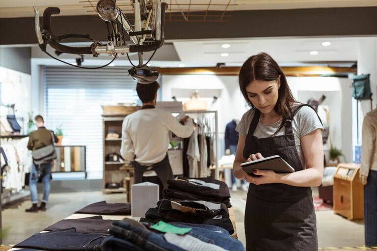 Female owner using digital tablet while coworker arranging clothes on rack at store Female owner using digital tablet while coworker arranging clothes on rack at store